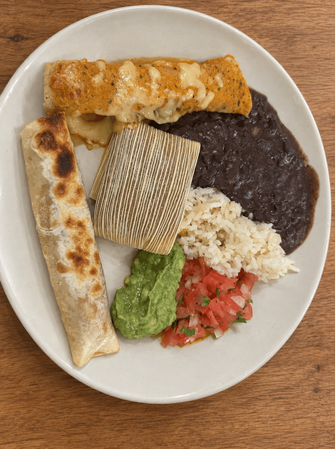 Plato combinado con enchilada en salsa roja cubierta de queso, burrito dorado de tortilla de harina, tamal en hoja de maíz, frijoles negros refritos, arroz, guacamole y pico de gallo