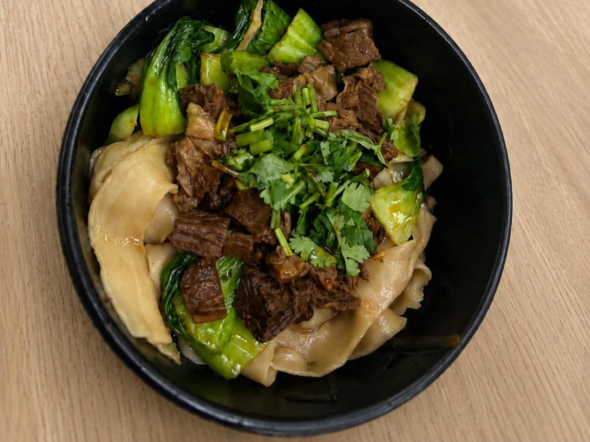Wide noodles with braised beef, bok choy, cilantro, and peppers in a black bowl