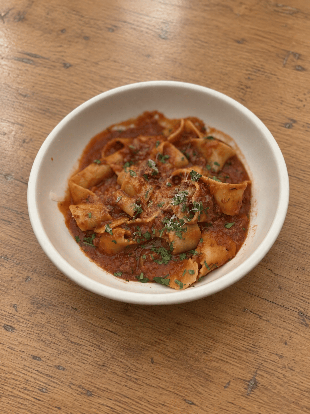 Wide pappardelle ribbons in Bolognese meat sauce with grated cheese and chopped herbs in a cream bowl.
