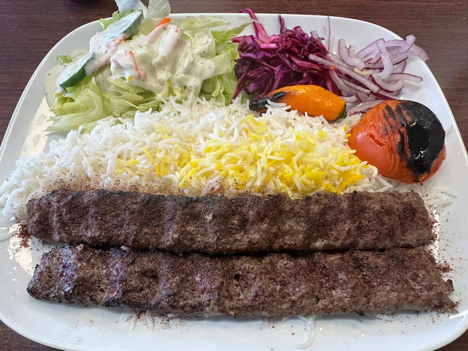 Two browned beef kebabs over white and saffron basmati rice with charred tomato, pepper, salad, and red onion