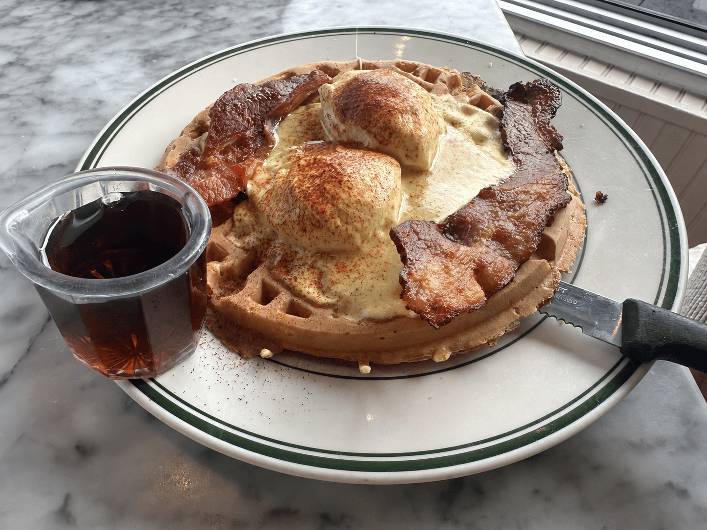 Bacon waffle topped with grilled ham, two poached eggs, hollandaise, and crisp bacon, with maple syrup on the side